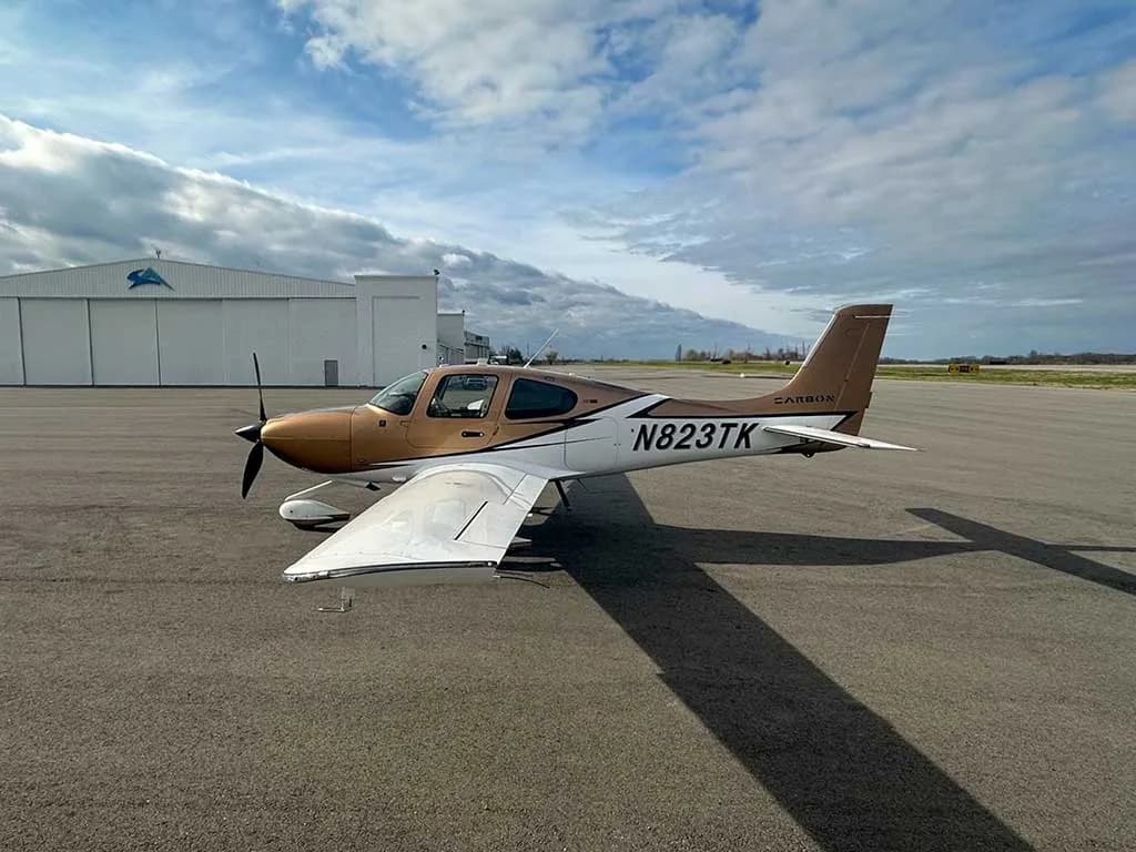 A Cirrus SR22 aircraft, N823TK, with a brown and white paint scheme parked on an airfield next to a hangar.