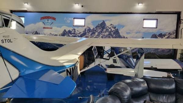 The rear-left side of a white and blue Maule aircraft, N1008W, parked in a hangar with a mountain mural and banner.