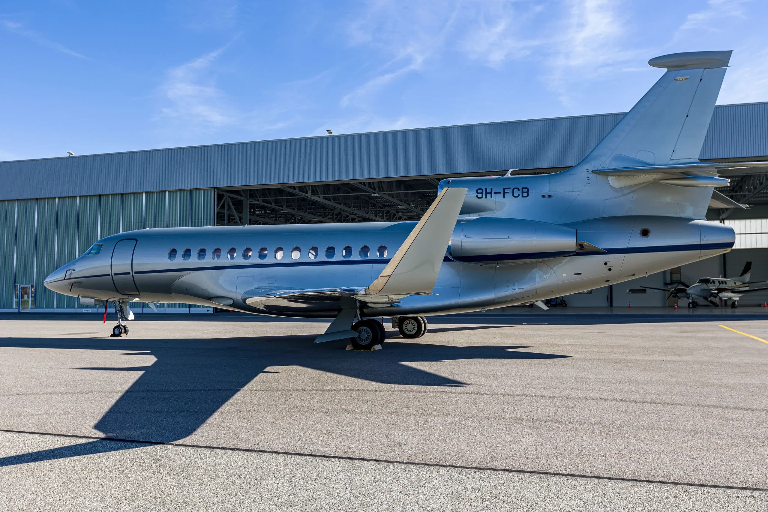 A silver Dassault Falcon 900EX business jet with blue stripes parked on an airfield, viewed from the rear-right side.