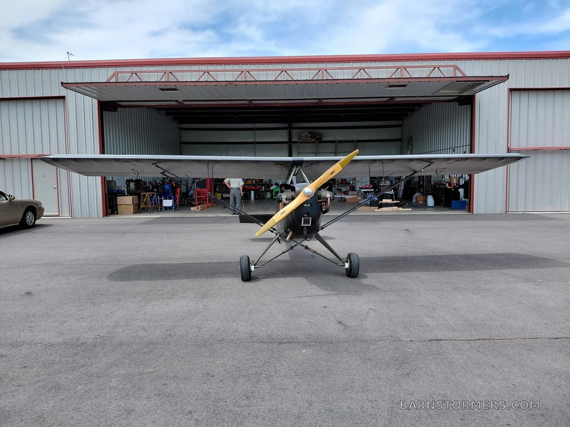 A dark green single-engine, high-wing aircraft with a wooden propeller, viewed head-on in front of an open hangar.