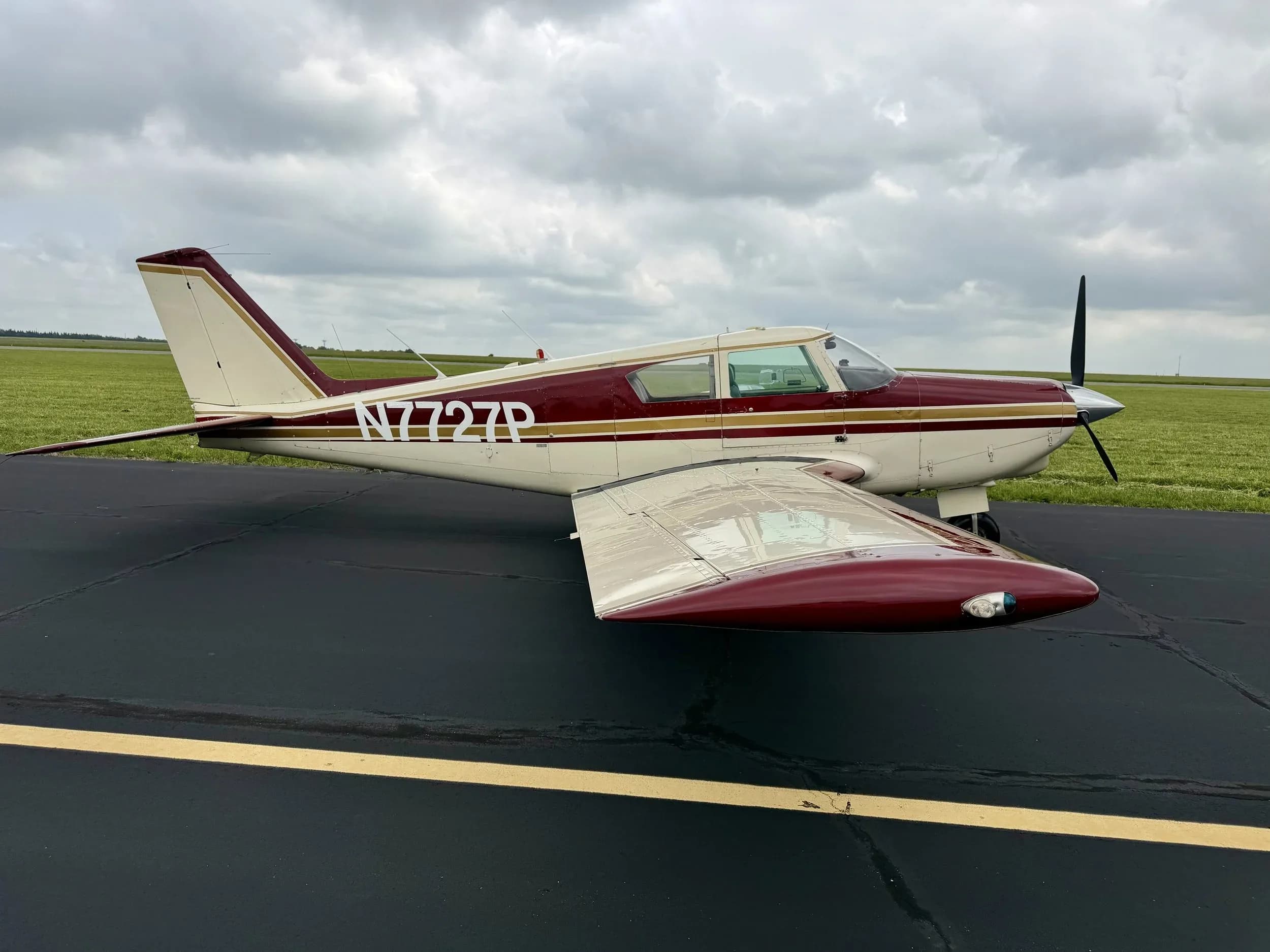 Side view of a maroon and white Piper Comanche N7727P with its registration N7727P visible on the fuselage.