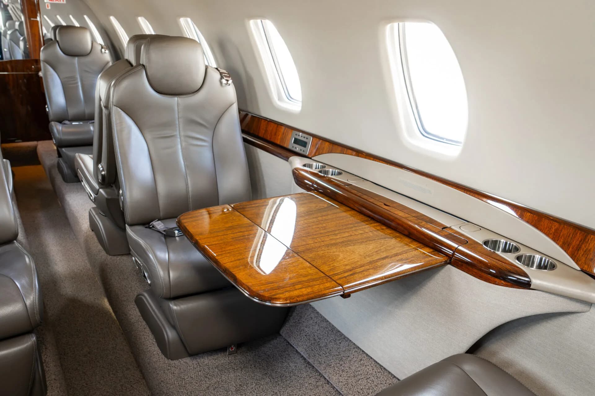 A close-up of a polished wooden fold-out table extended from the sidewall next to a tan leather seat in a business jet.