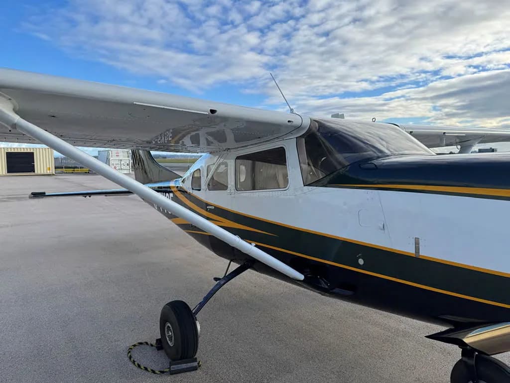A close-up view of the front fuselage and wing of the dark green and white Cessna 182, showing the propeller and landing gear.