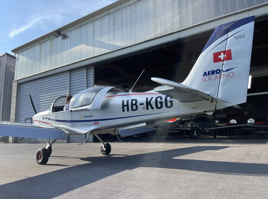 The tail section of a silver Sonaca 200 aircraft with Swiss flag and HB-KGG registration is visible outside a hangar.