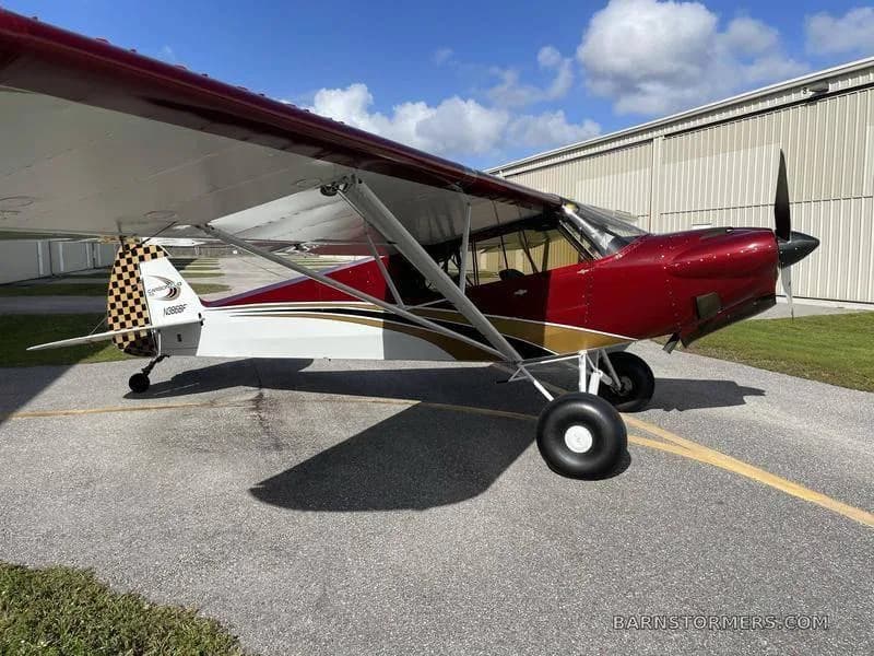 A red and white CubCrafters aircraft with gold trim, registration N386BF, parked on an asphalt apron in front of a hangar.