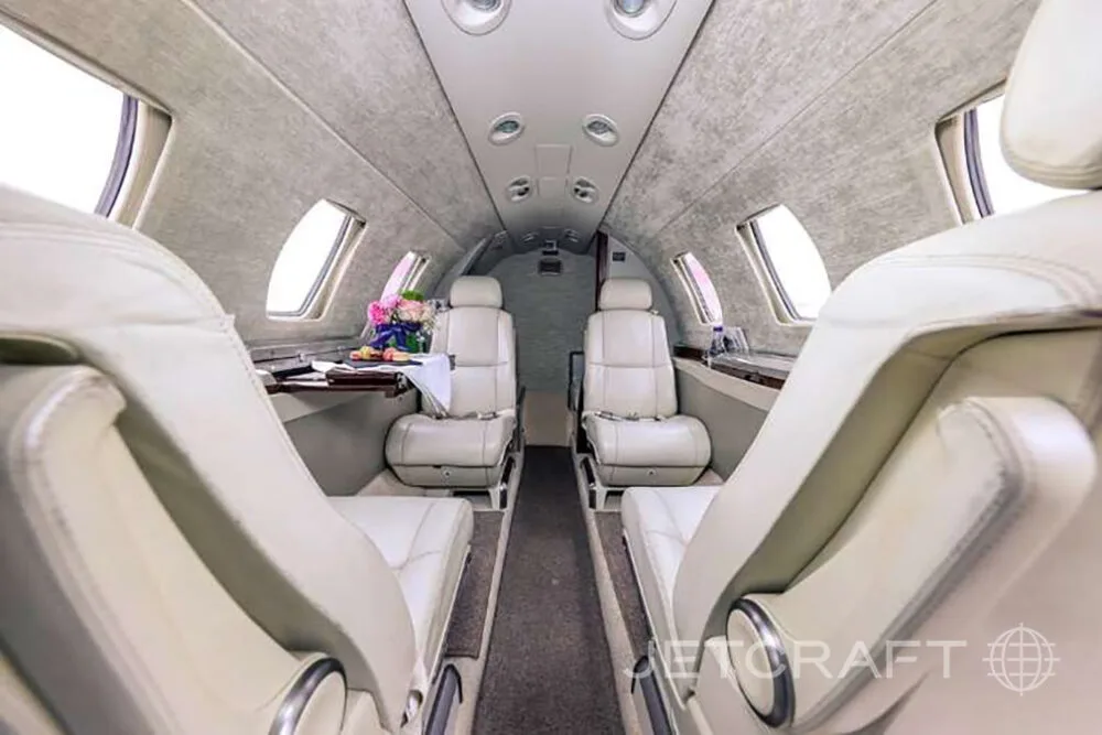 A view down the aisle of a private jet's cabin, showing light-colored leather seats and a small table with flowers.