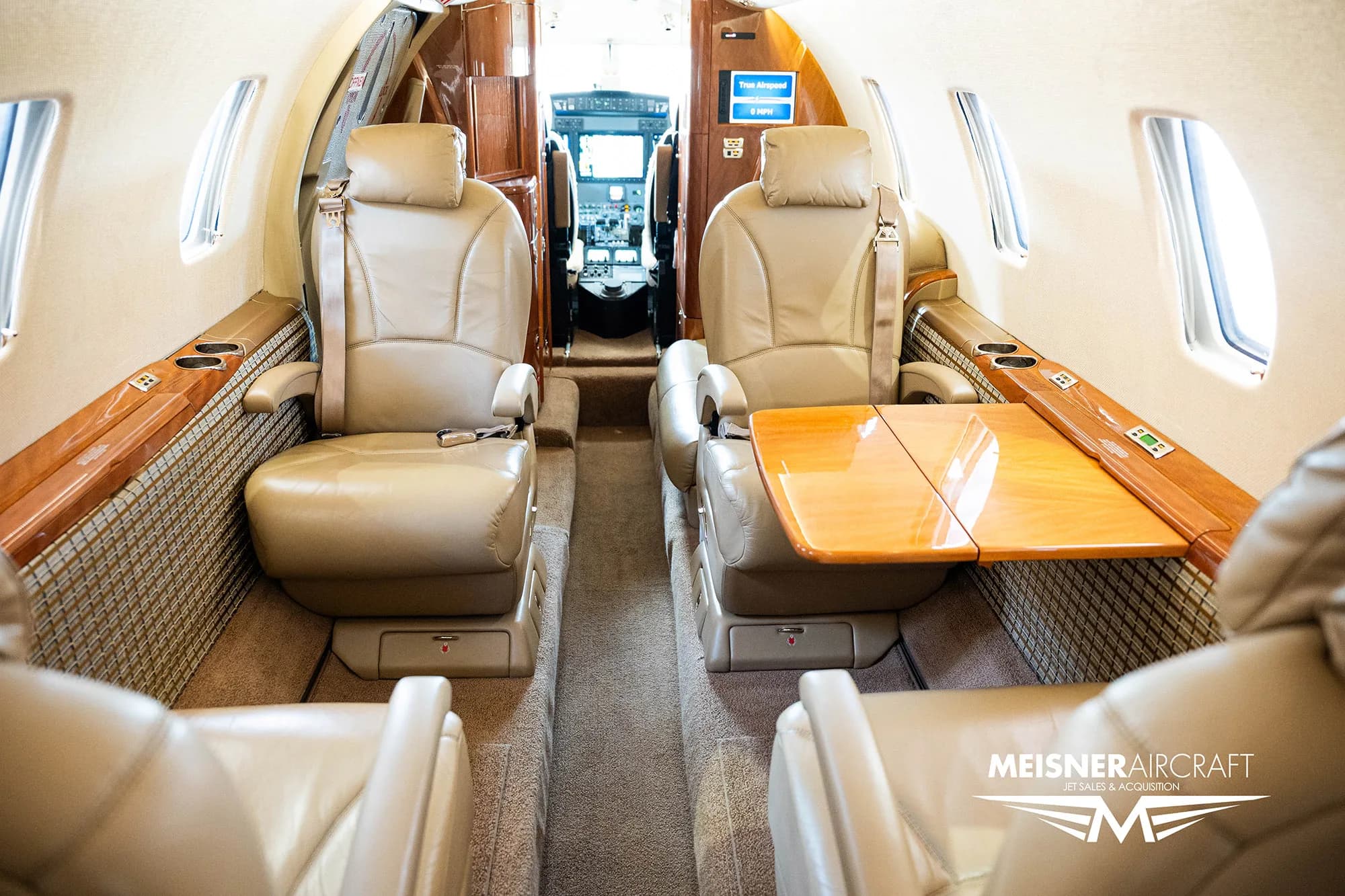 The interior cabin of a business jet with tan leather seats and a wooden table deployed between them, looking towards the cockpit, with a 'Meisner Aircraft' logo.