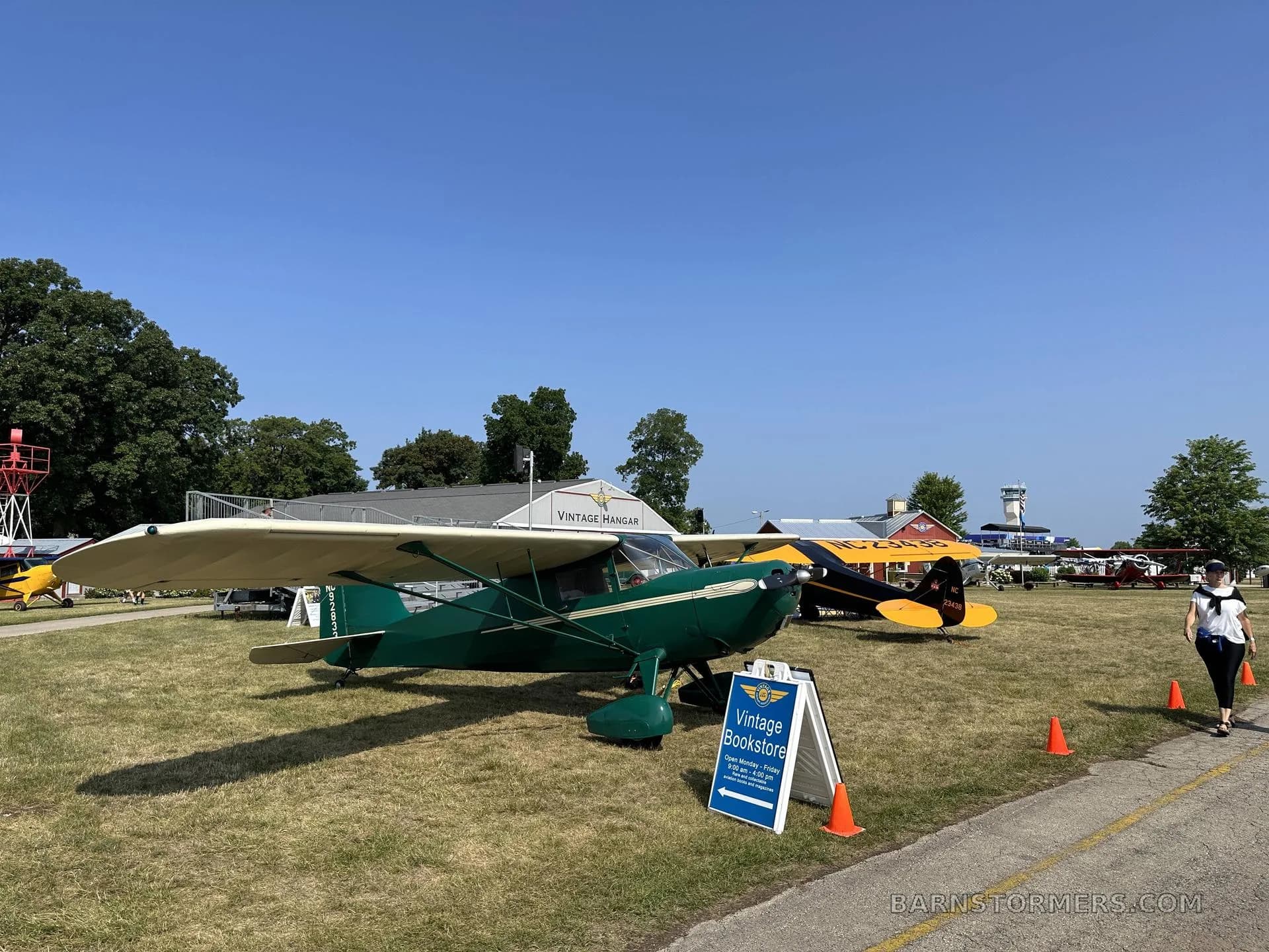 A green and cream vintage aircraft with tail number NC92833 is parked on grass at an airfield, with other aircraft and buildings in the background.