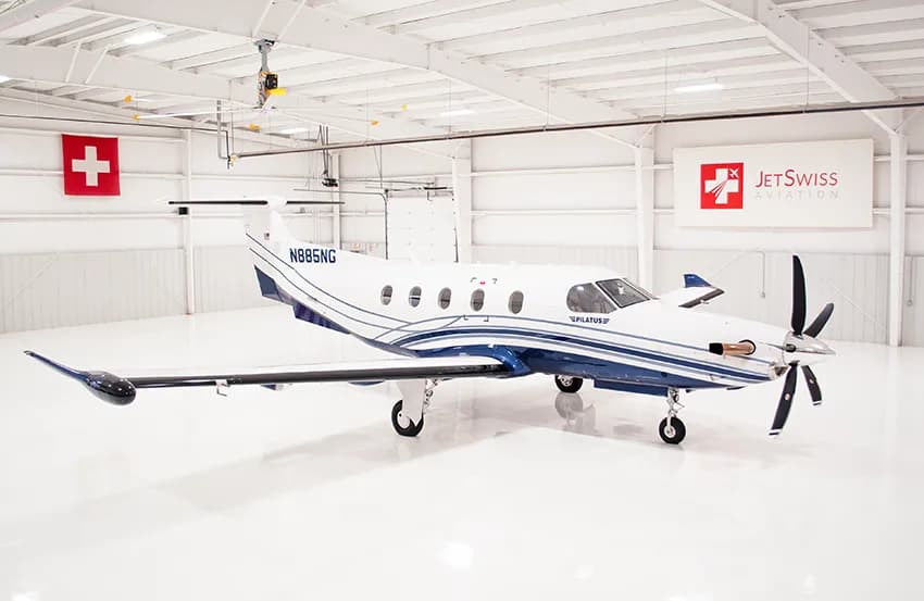 A white Pilatus PC-12 with blue and silver stripes is parked inside a brightly lit white hangar.