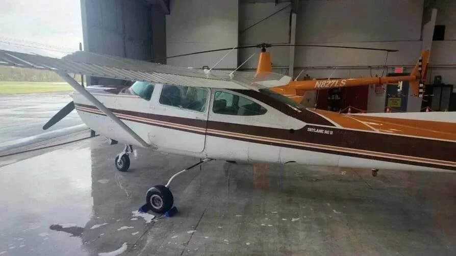 A white and brown Cessna Skylane RG II aircraft parked inside a hangar, with an orange helicopter visible behind it.