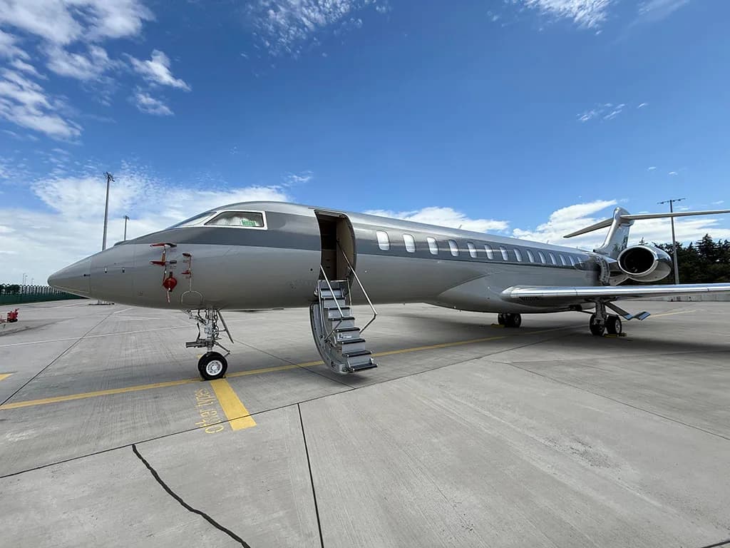 A grey and silver private jet with its door open and stairs extended, parked on an airport tarmac under a blue sky.