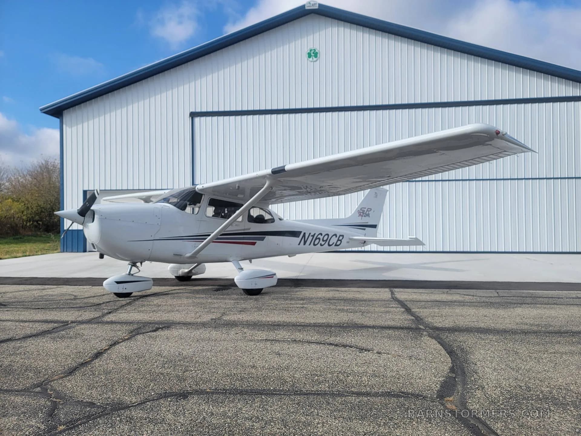 A white Cessna Skyhawk N169CB with black and red stripes is parked on a cracked asphalt apron next to a white hangar.