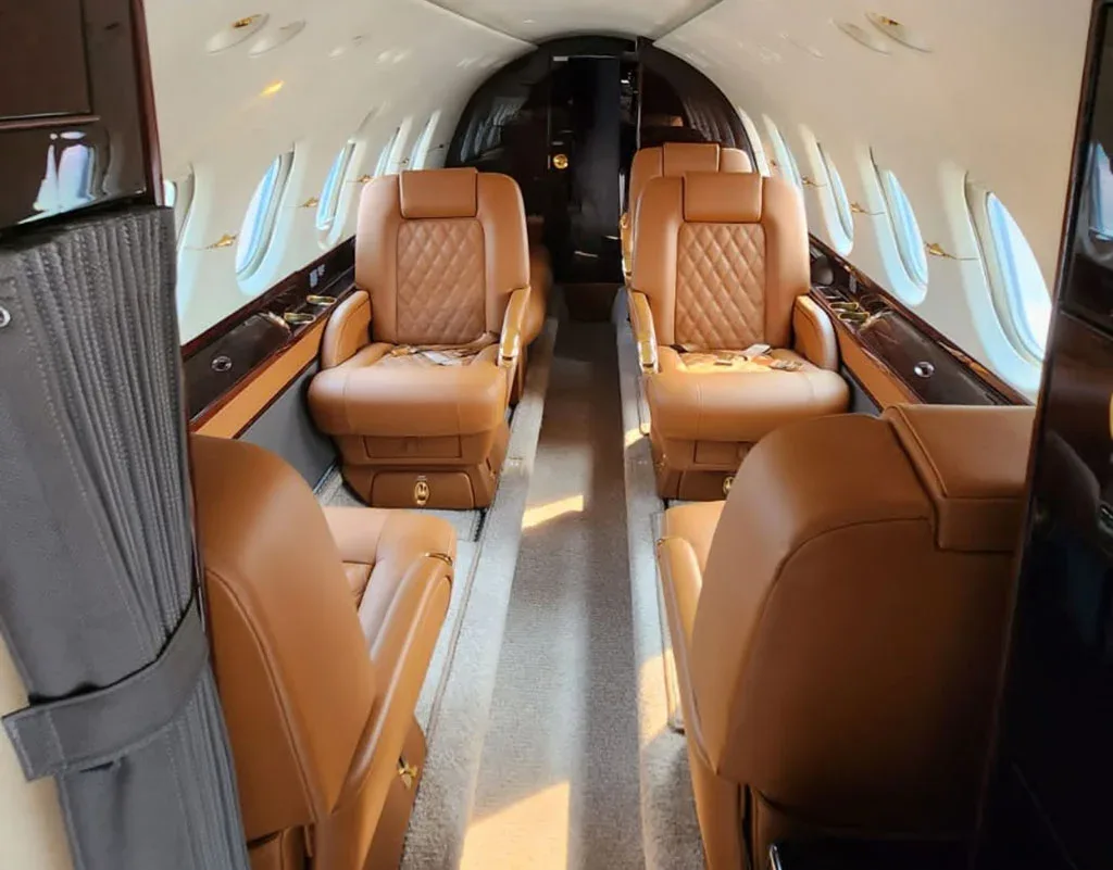 A view of the business jet cabin from the rear, showing multiple rows of brown leather seats and a carpeted aisle.