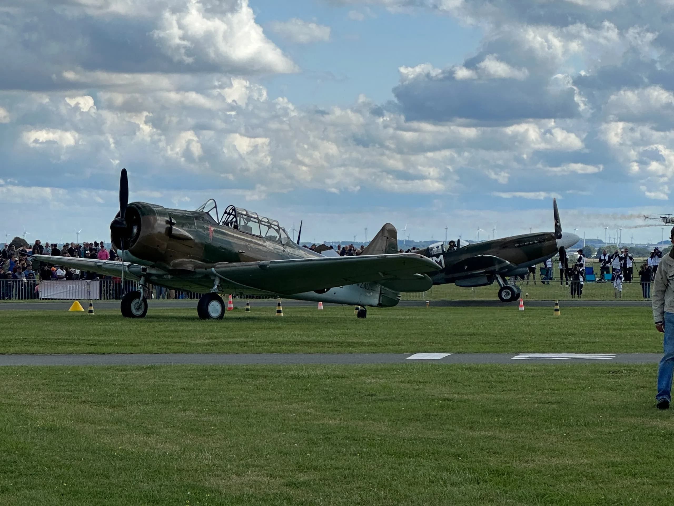 Two vintage military aircraft, including the F-AZAU North American T-6 Texan, parked on a tarmac with a crowd in the distance.
