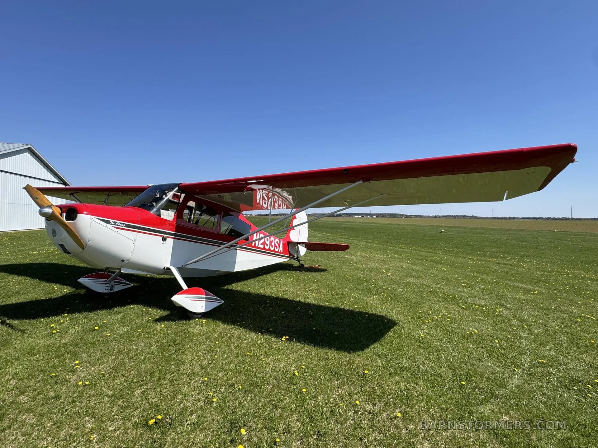 A red and white single-engine aircraft with tail number N293SA parked on a grassy field next to a hangar.