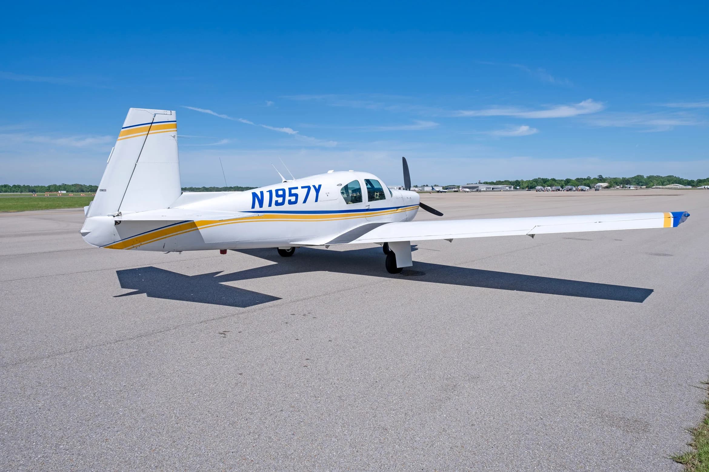 A rear-side view of a white single-engine propeller aircraft with blue and yellow stripes, registration N1957Y, parked on an asphalt apron.