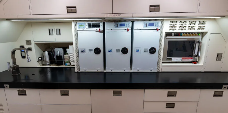 A well-equipped aircraft galley features a long black countertop, multiple appliances, and white storage cabinets.