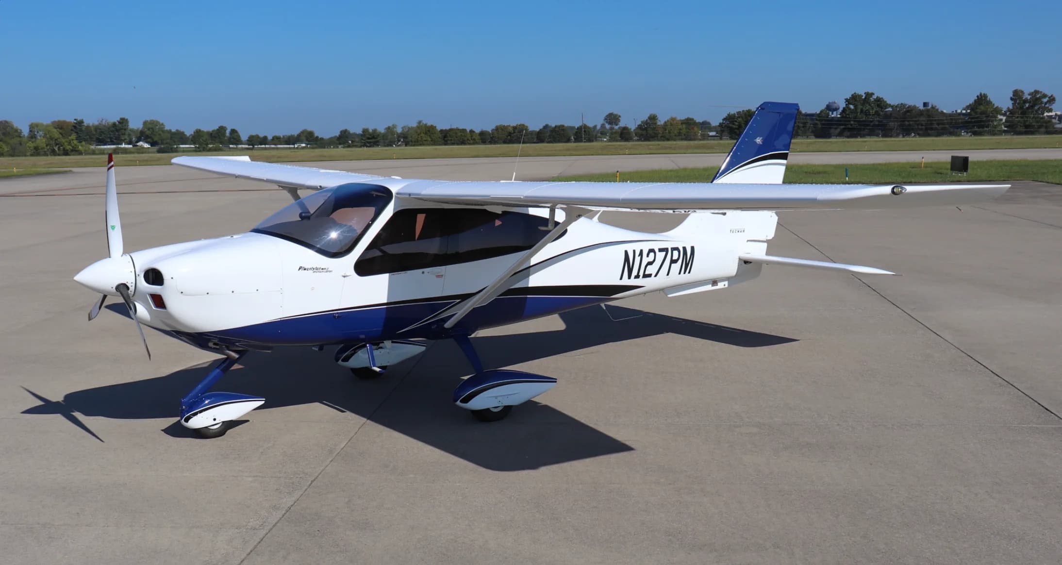 An elevated view of the white and blue Tecnam P2010 aircraft, registration N127PM, parked on an airfield.