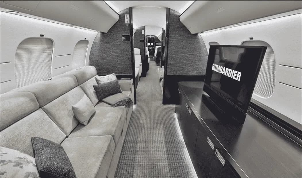 A luxurious aircraft cabin interior with a large sofa on the left and a TV displaying "BOMBARDIER" on the right.