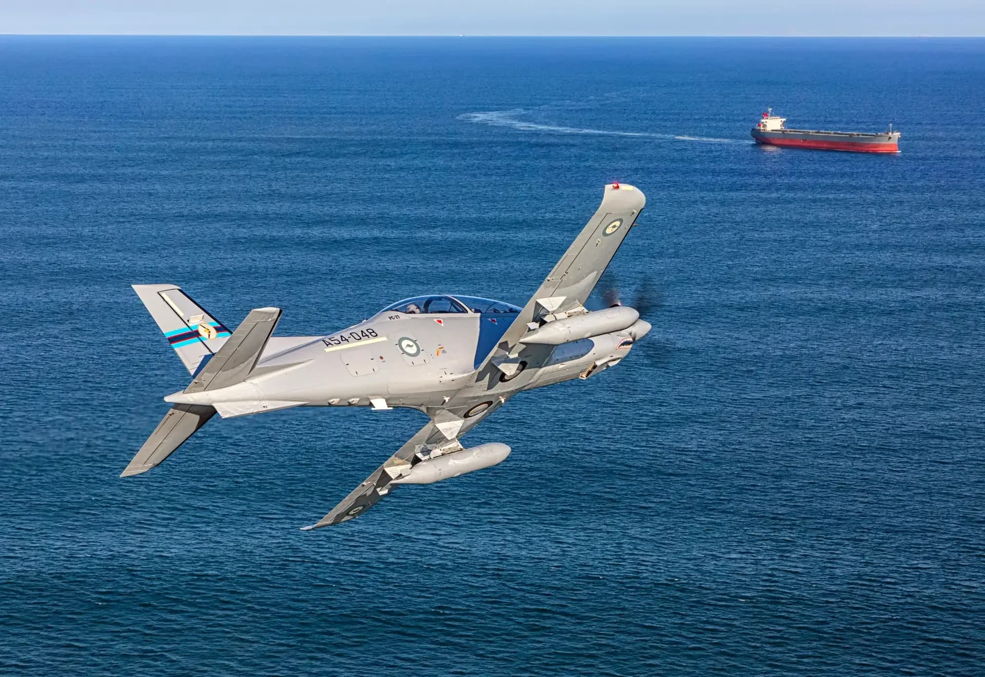 A gray Pilatus PC-21 aircraft flying over the ocean with a ship in the background.