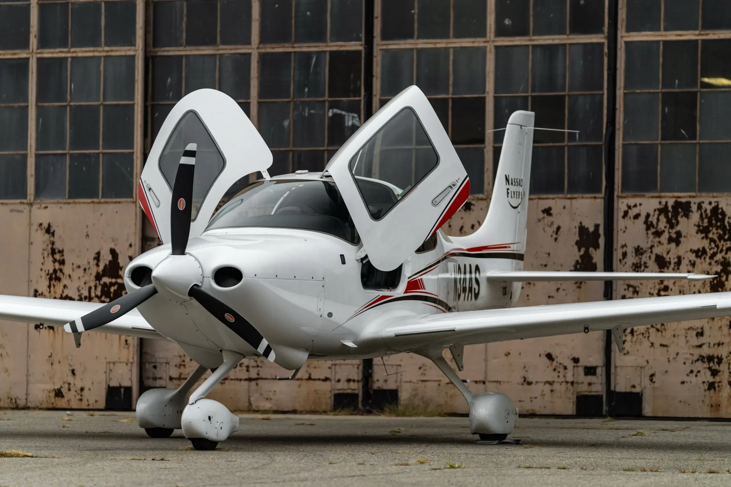 A white Cirrus SR22 aircraft with red and black stripes, registration N34AS, parked with its gull-wing doors open in front of an old hangar.