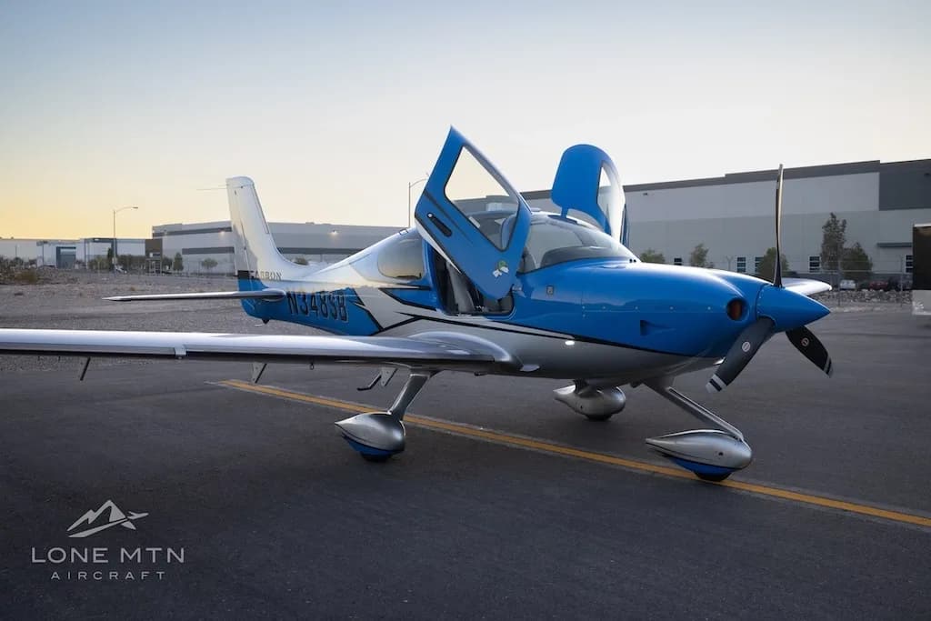 A blue and silver Cirrus SR22 aircraft with open doors parked on an airfield with hangars in the background at sunset.