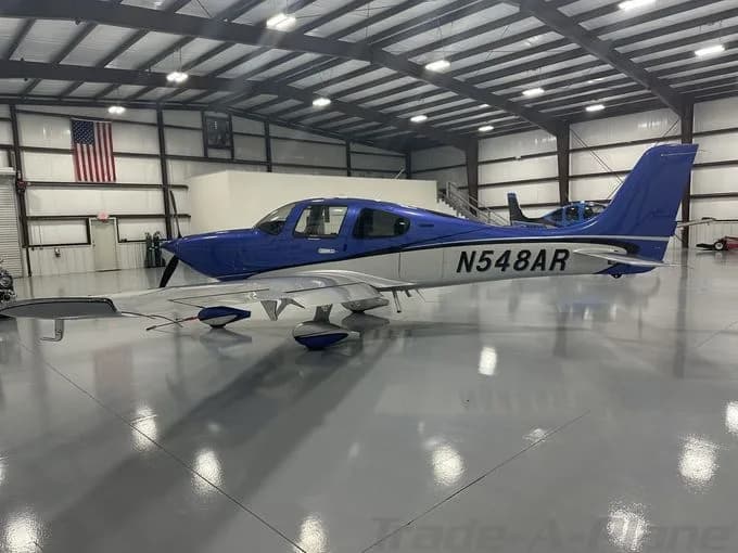 A blue and white single-engine aircraft, N548AR, is parked inside a brightly lit hangar with an American flag on the wall.
