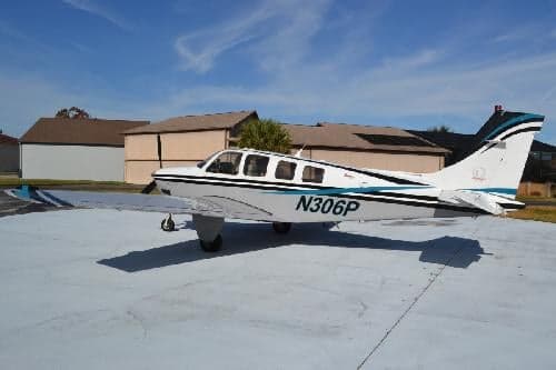 A Beechcraft Bonanza aircraft with a white and blue paint scheme parked on a concrete surface with buildings in the background.