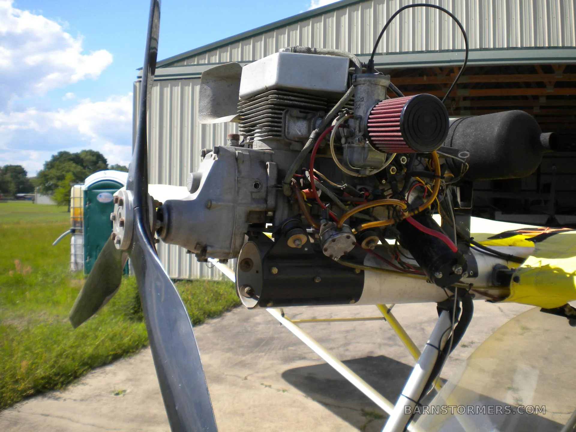 A close-up view of the ultralight aircraft's engine, propeller, and air filter, with a hangar in the background.
