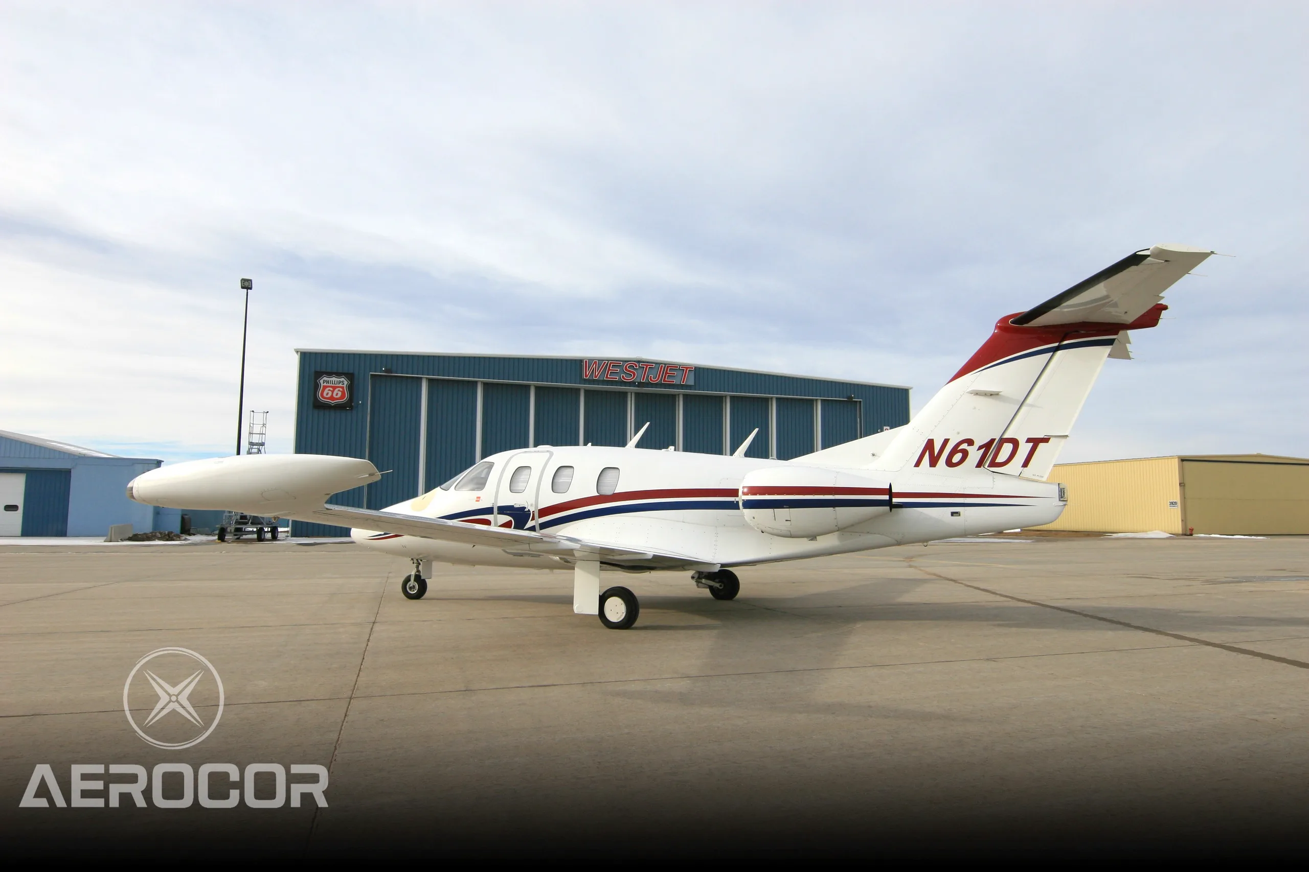 A white Eclipse 500 jet with red and blue stripes parked on an airport tarmac in front of a Westjet hangar.
