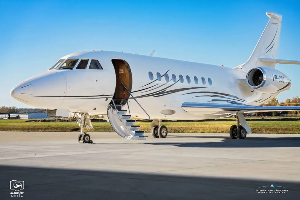 A white Dassault Falcon business jet with black and silver livery, registration VP-CDJ, parked on an tarmac with its main door open.