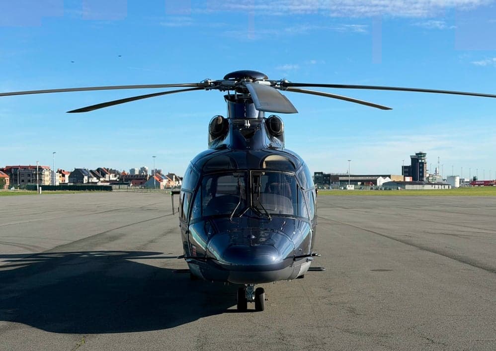 A dark blue helicopter is parked on an airfield, viewed from the front, with buildings in the distance.