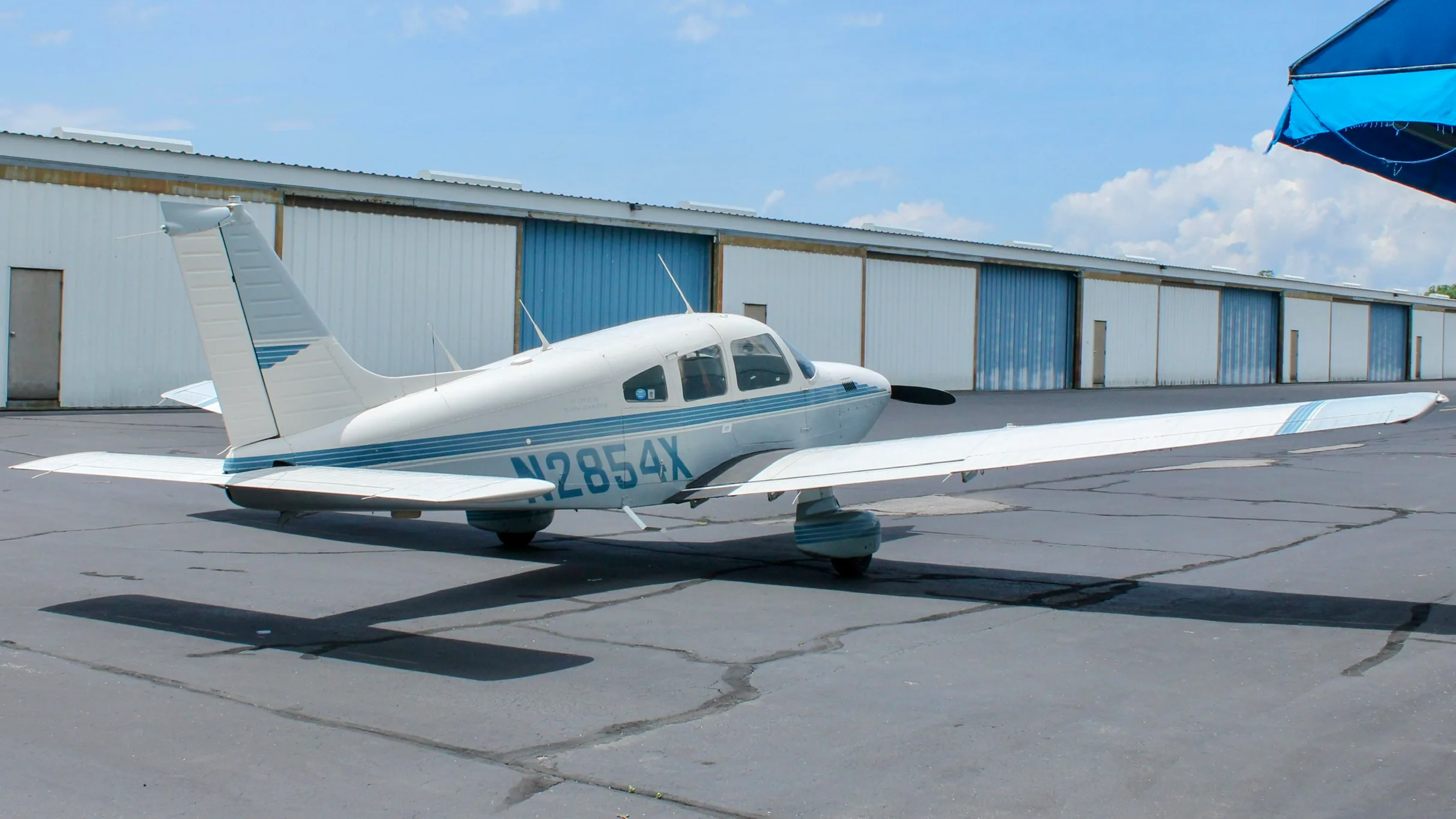 A side view of a white and blue Piper Cherokee aircraft, registration N2854X, parked on an asphalt ramp with hangars in the background.