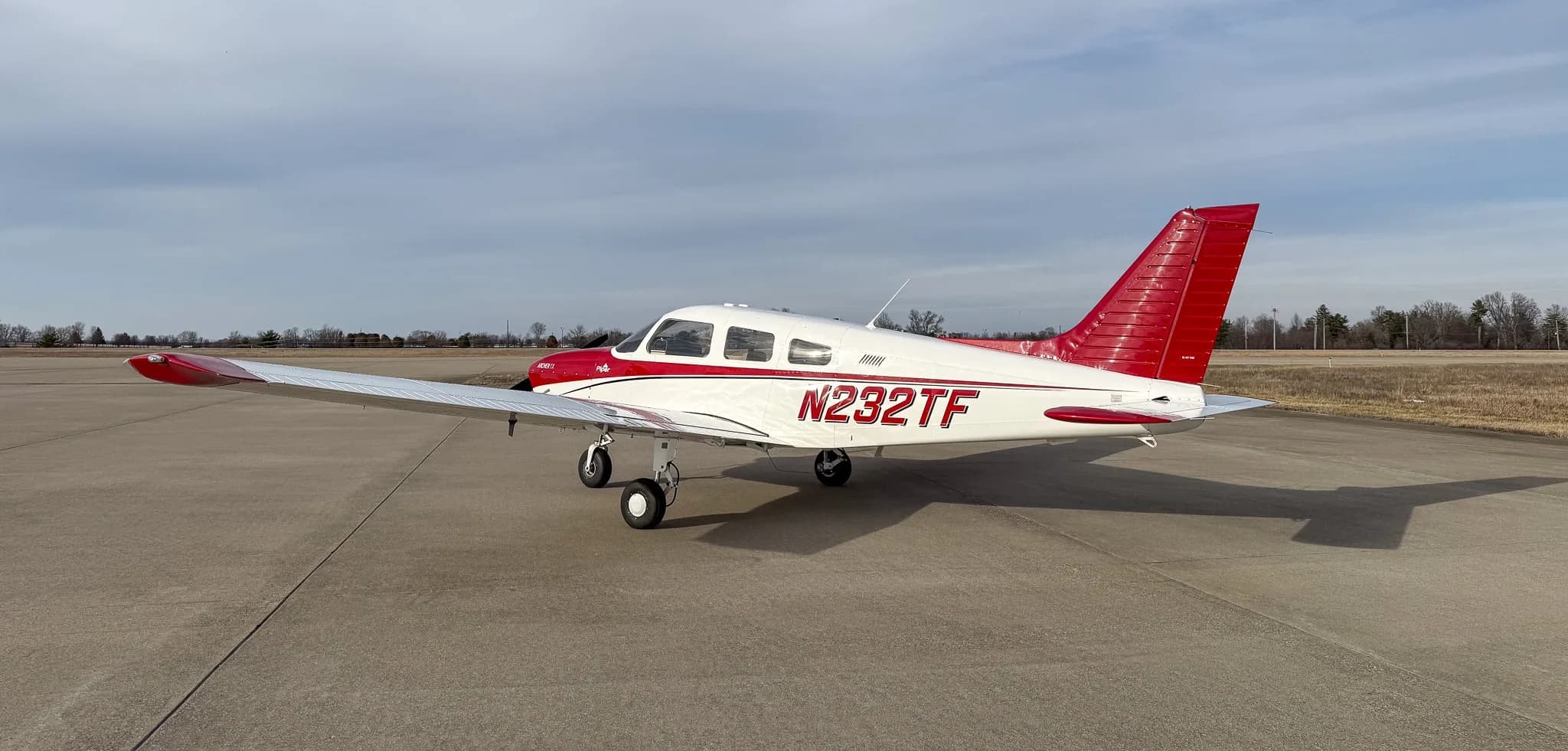 A side view of a white and red Piper Archer TX, N232TF, parked on an airfield with its shadow cast on the ground.
