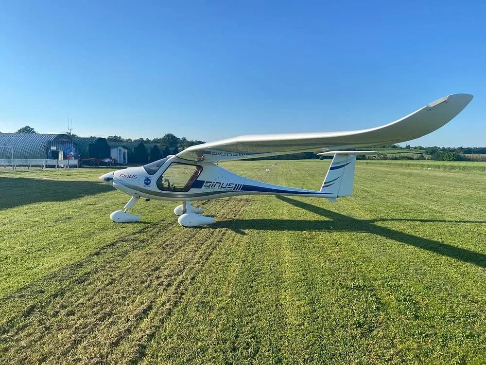 A white Pipistrel Sinus aircraft with blue and green tail stripes, viewed from the side on a bright green airfield.