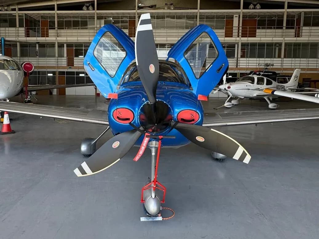 A head-on view of a blue and silver Cirrus SR22 aircraft with its gull-wing doors open, parked in a hangar.