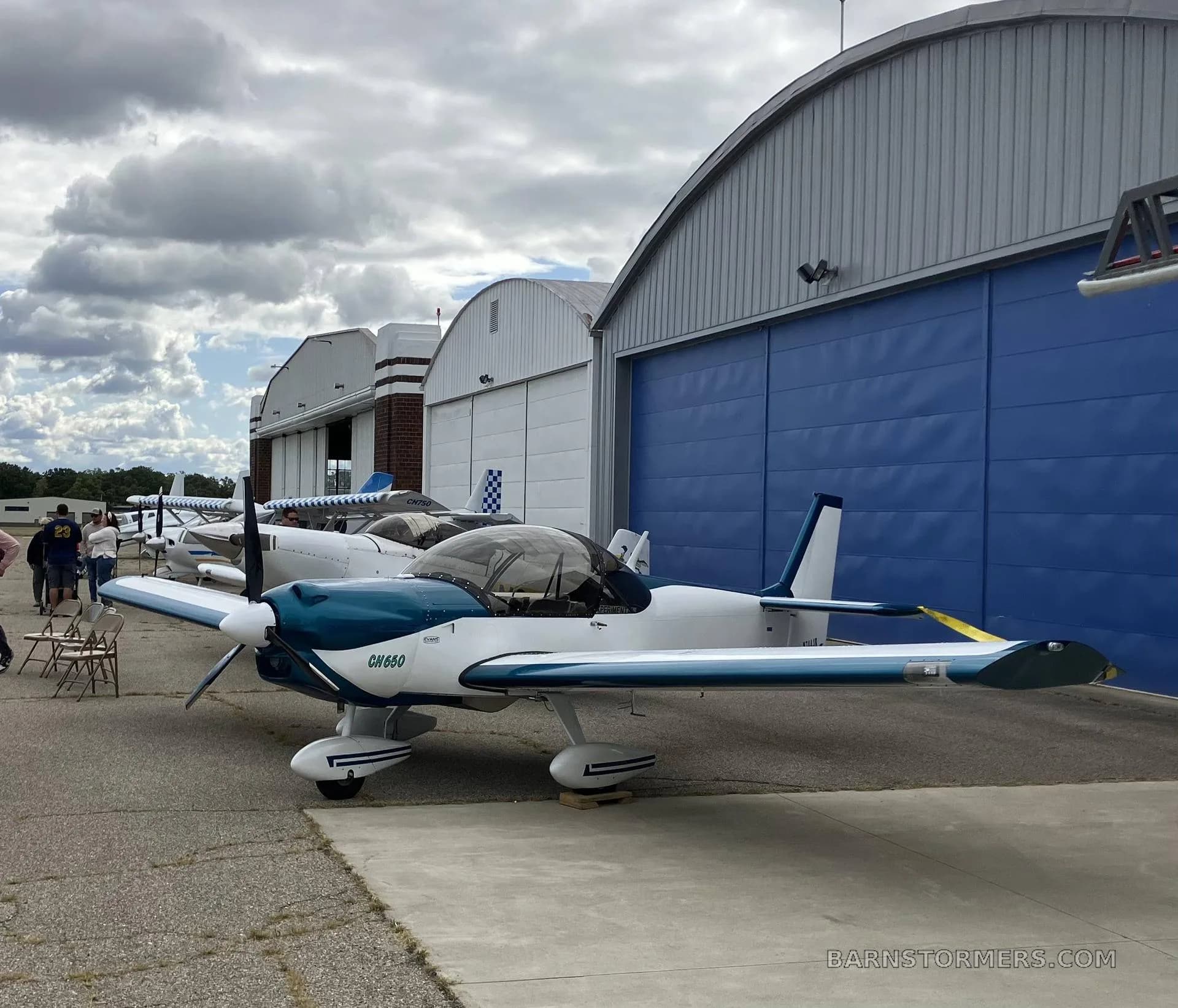 A white and blue Zenith CH650 aircraft with registration N744JK is parked on an airfield tarmac next to blue hangars under a cloudy sky.