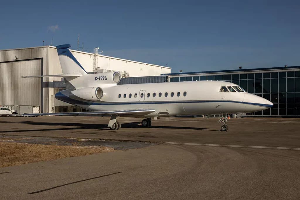 A white and blue Dassault Falcon 900EX business jet with registration C-FPFS parked on an airfield next to hangars.