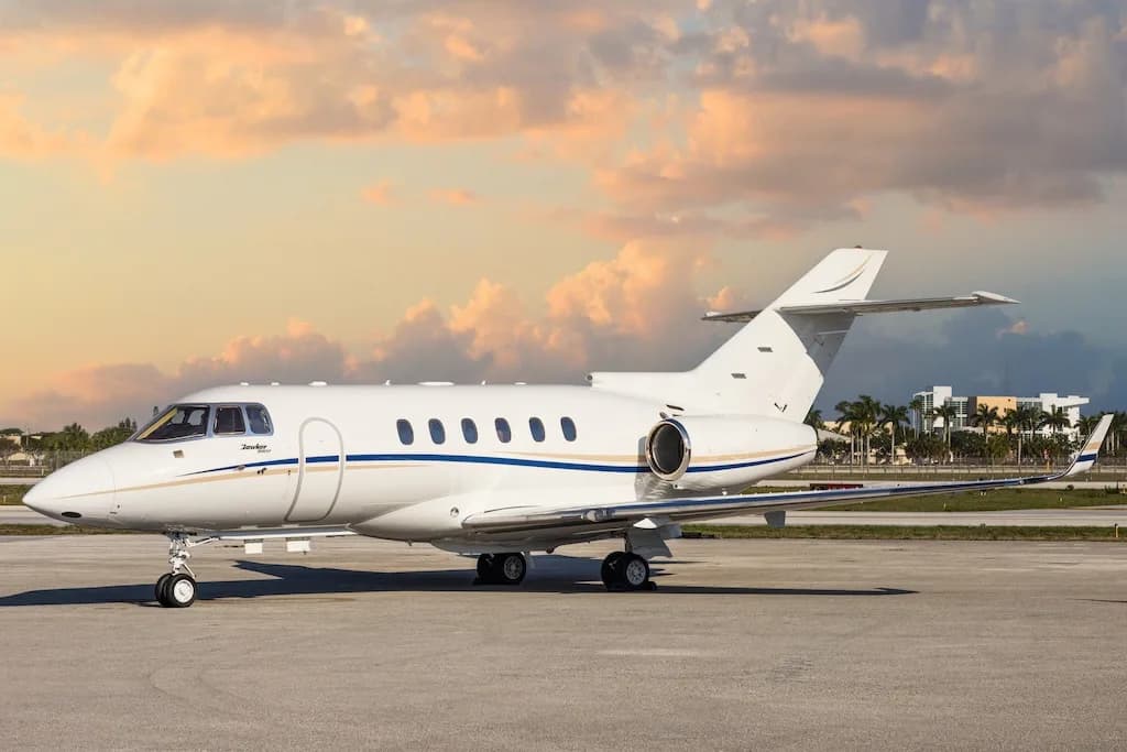 A white Hawker business jet with blue and gold stripes is parked on an airport tarmac at sunset.