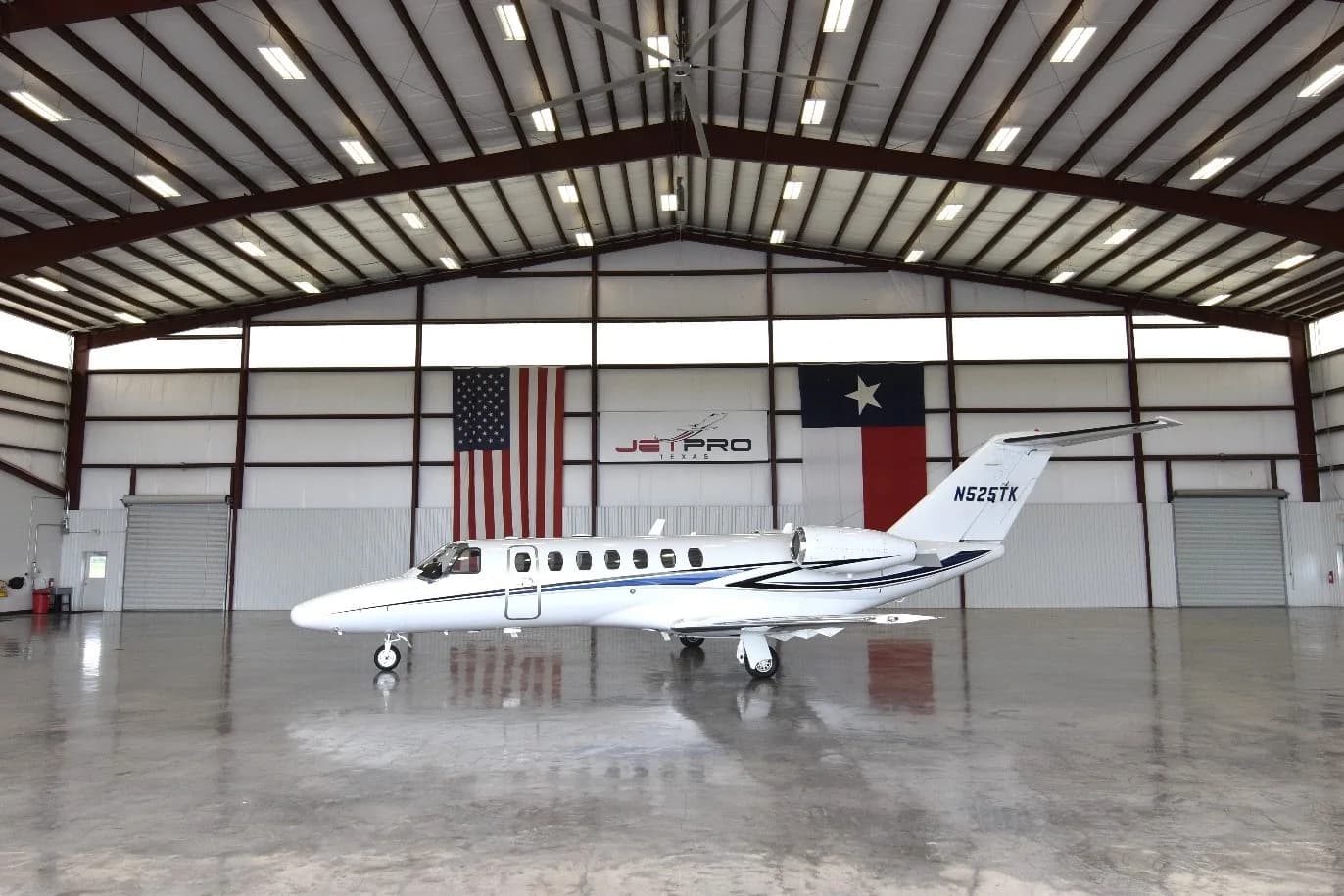 A white Cessna Citation CJ3 private jet with blue and black stripes, registration N525TK, parked in a hangar with US and Texas flags.