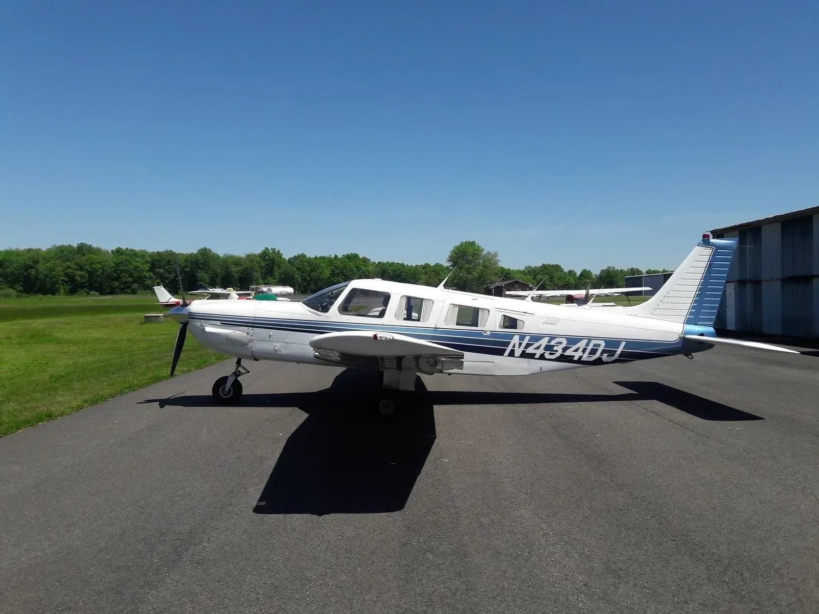 A side view of the white Piper PA-32R-300 Cherokee Lance, registration N434DJ, with blue stripes, parked on an airfield.