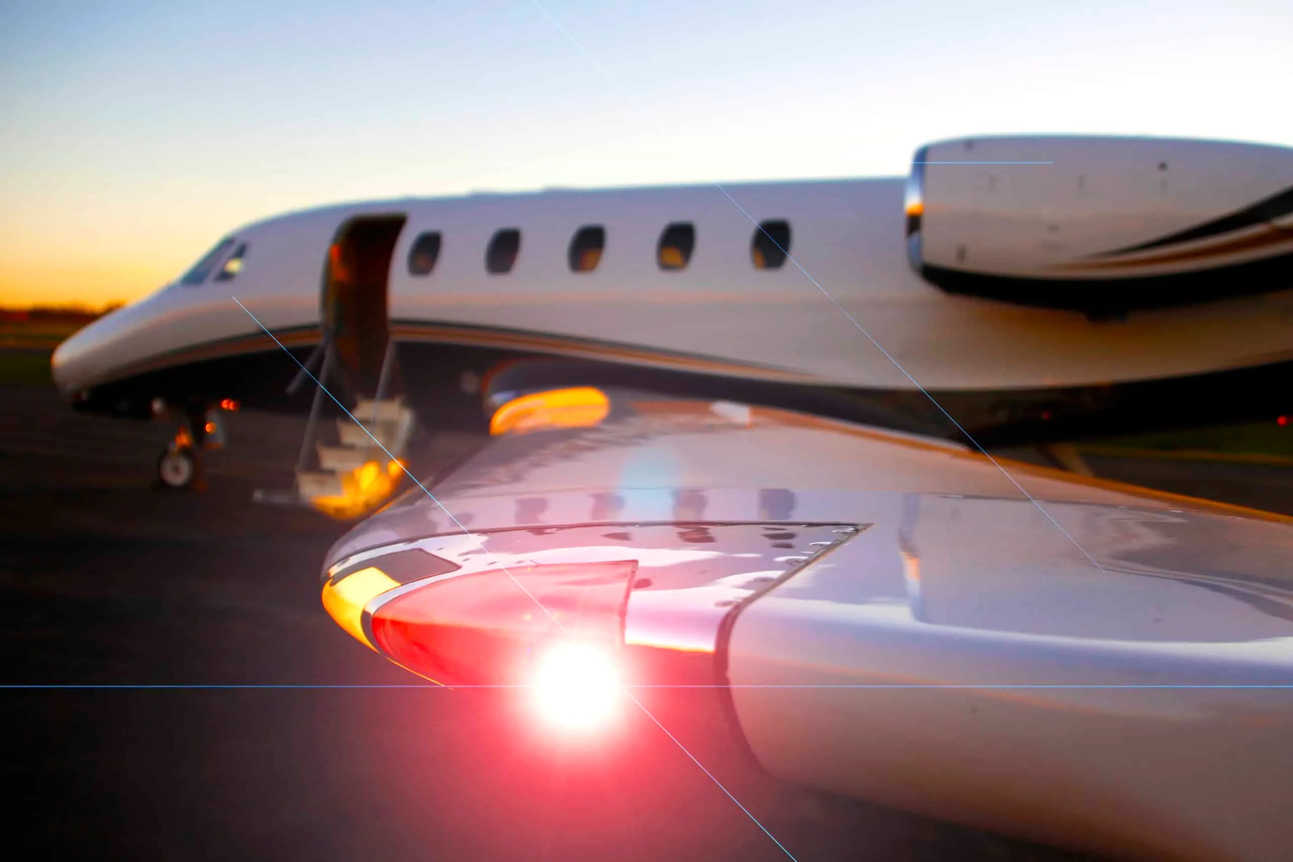 A close-up view of the wingtip and fuselage of a white private jet with its door open, showing a bright red navigation light at sunset.