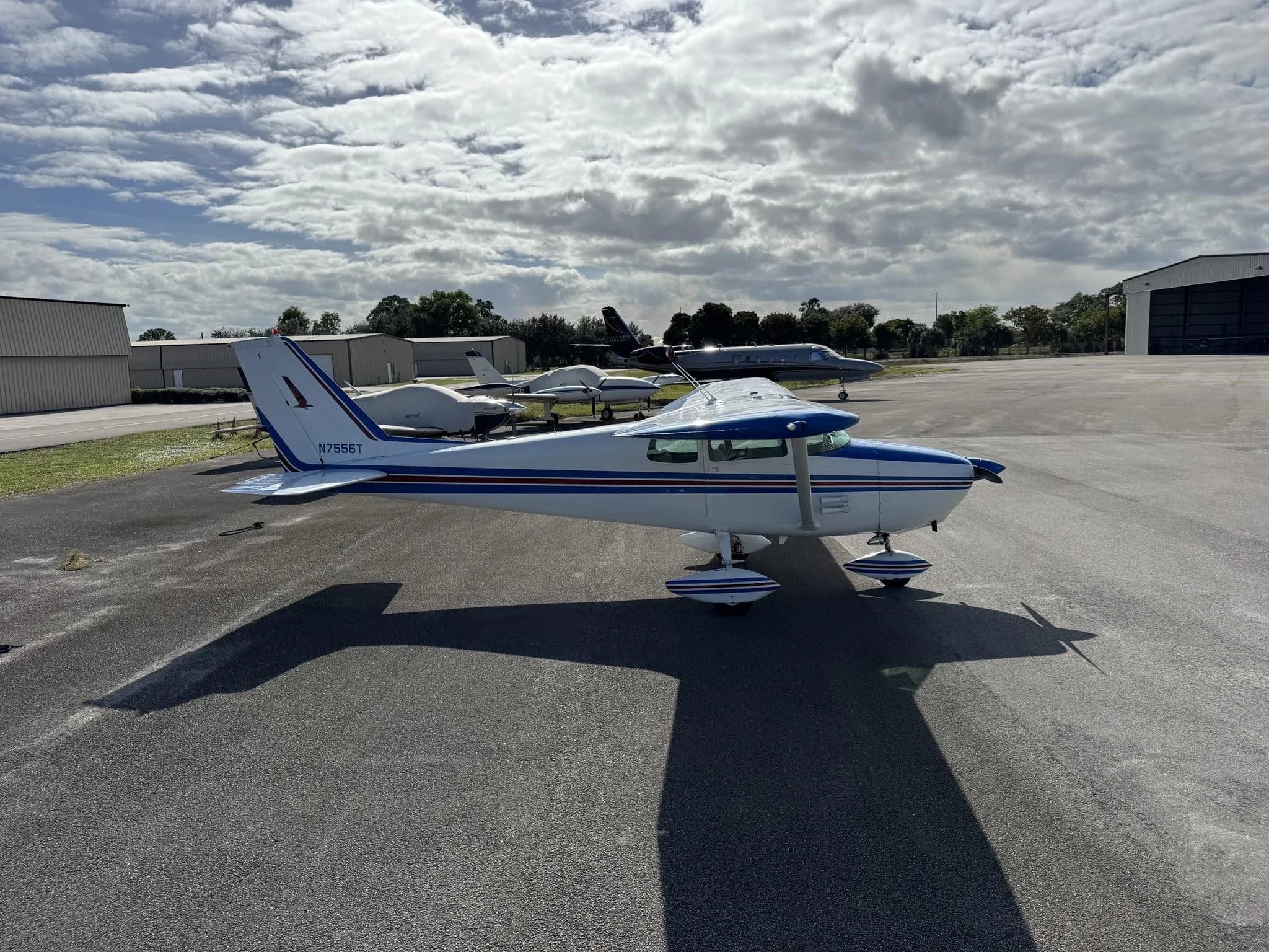 Side view of a white and blue Cessna 172 (N7556T) parked on an airfield with other aircraft in the distance.