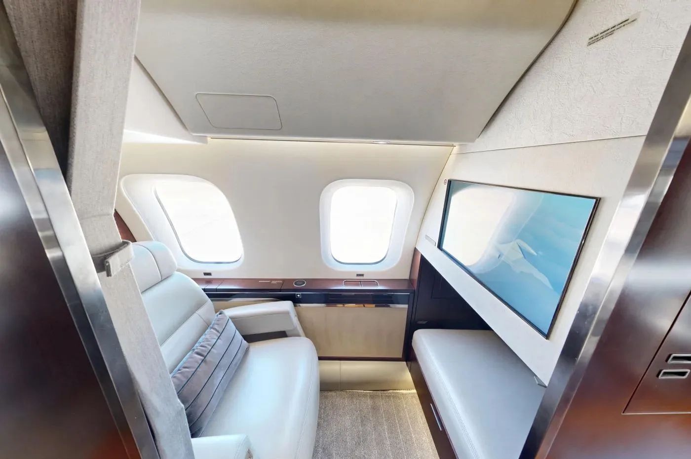 A private jet cabin lounge area with a cream leather seat, a side bench, and a large wall-mounted screen.