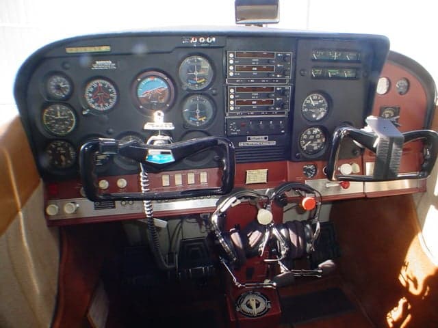 The cockpit of a small aircraft, featuring a dark instrument panel with various gauges and two control yokes.
