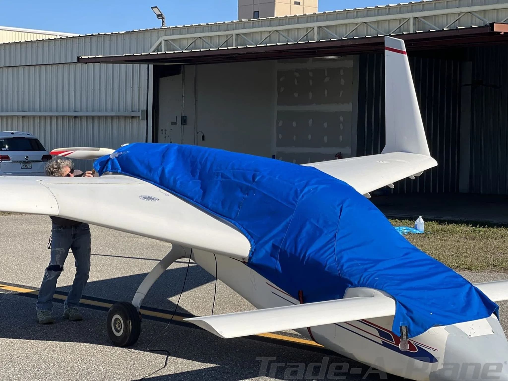 A person covers the fuselage of a white experimental aircraft with a blue tarp, parked on an airfield next to a hangar.