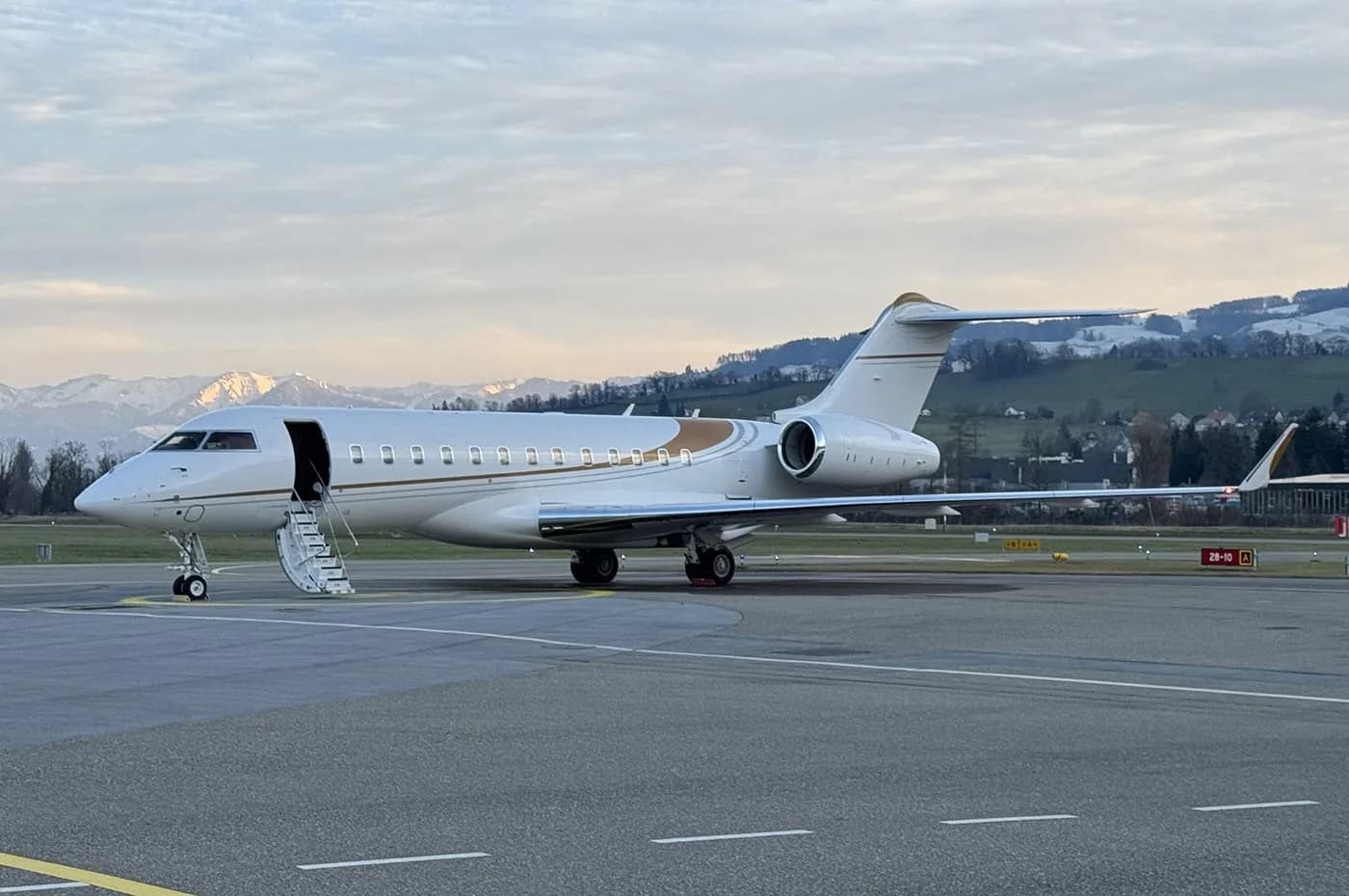 A white and gold business jet, likely a Bombardier Global, is parked on an airfield with its door open and stairs extended, against a backdrop of mountains.