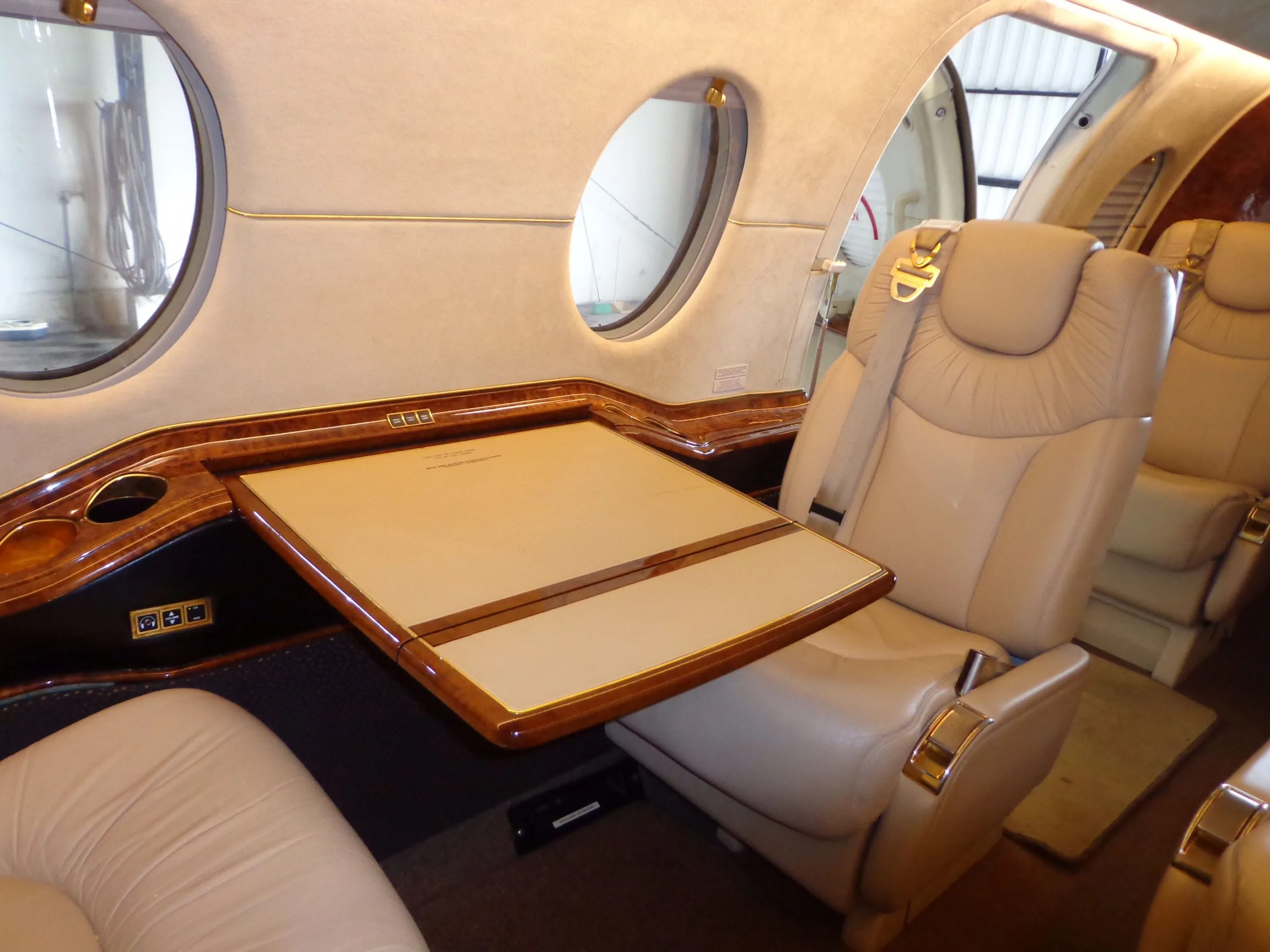 A private jet interior showing a beige leather seat and a fold-out wooden table next to a window.