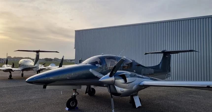 A dark blue and silver Diamond DA62 twin-engine aircraft parked on an apron in front of a hangar at sunset.