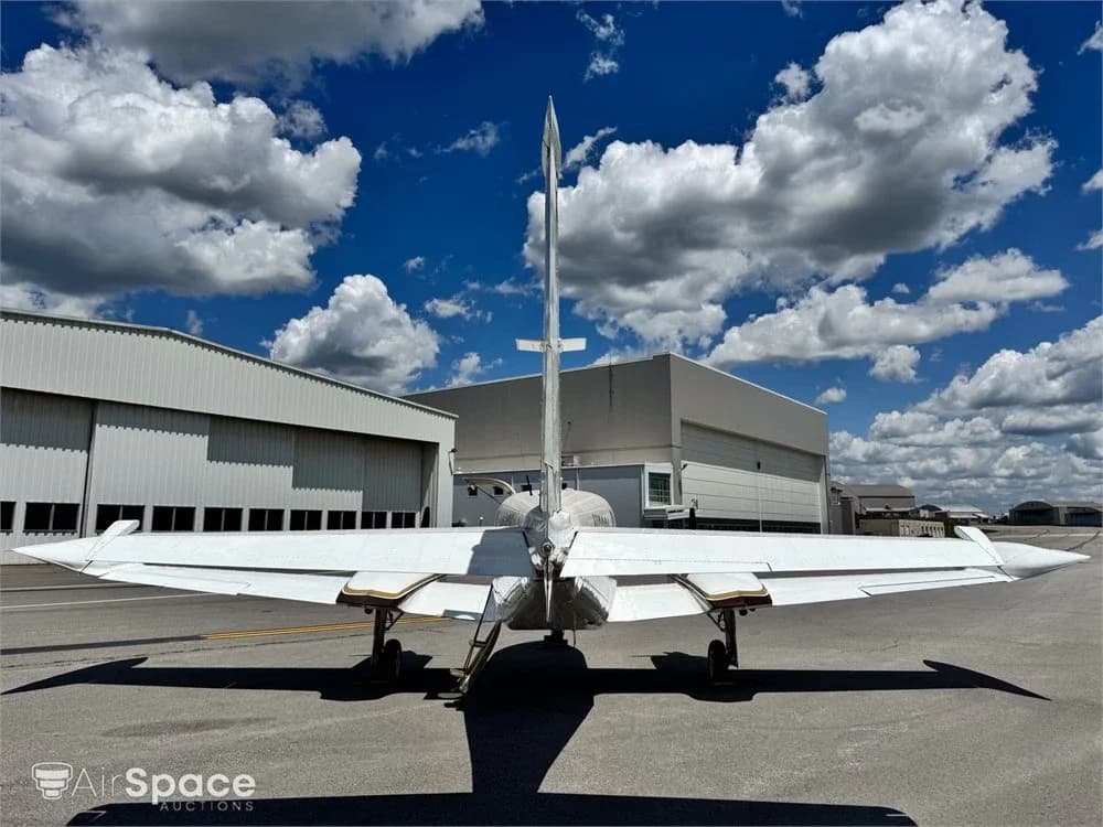 A rear view of a white twin-engine aircraft with gold and brown stripes parked on an asphalt ramp in front of hangars.